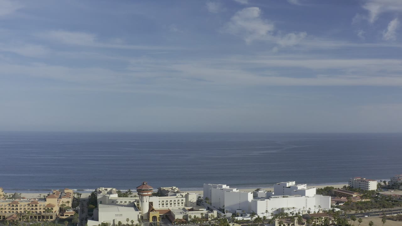 Opening Los Cabos aerial frame with blue ocean and pale sky above white coastal buildings, roads, palms, and no visible people.