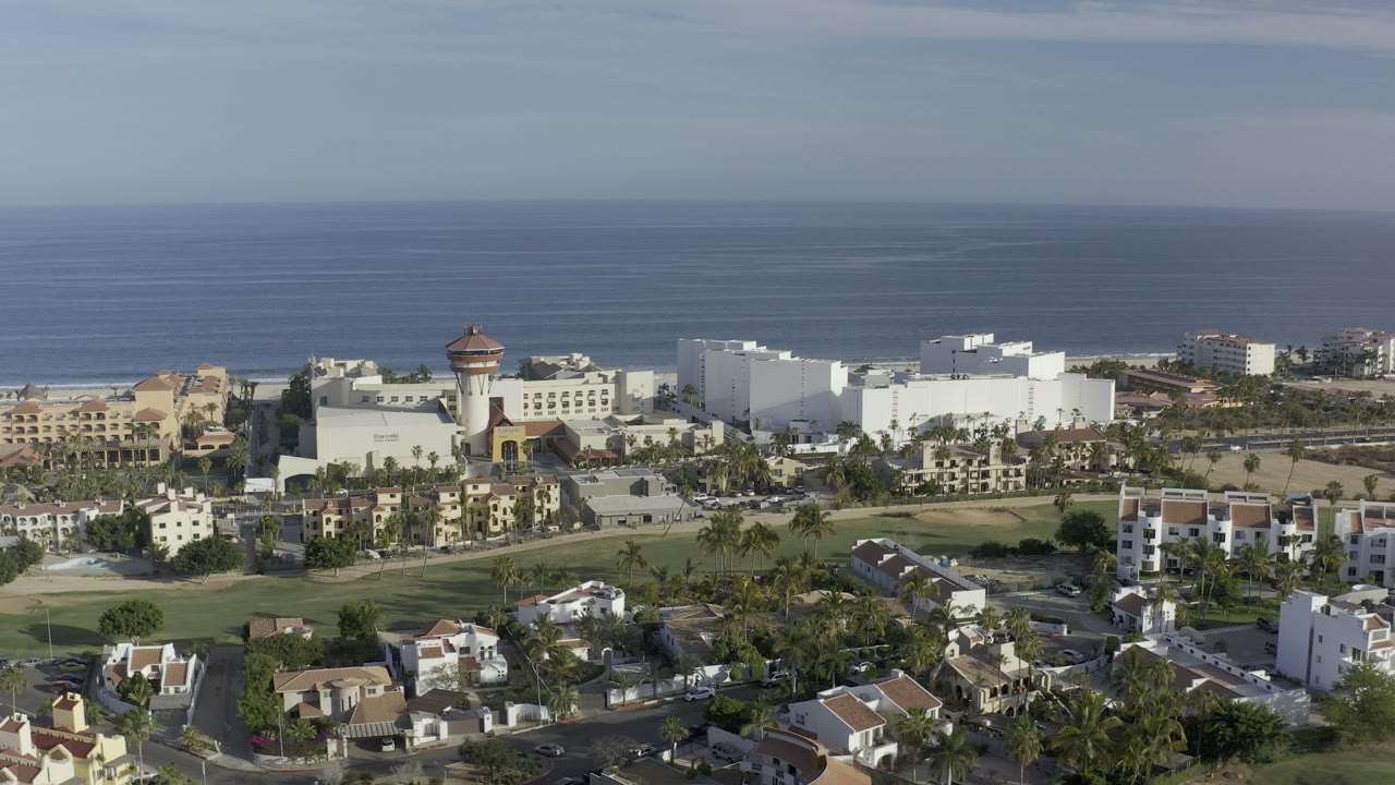 Wide aerial view in Los Cabos, Mexico, showing clustered homes, green lawns, white coastal buildings, and the ocean horizon in clear daylight, with no visible people.