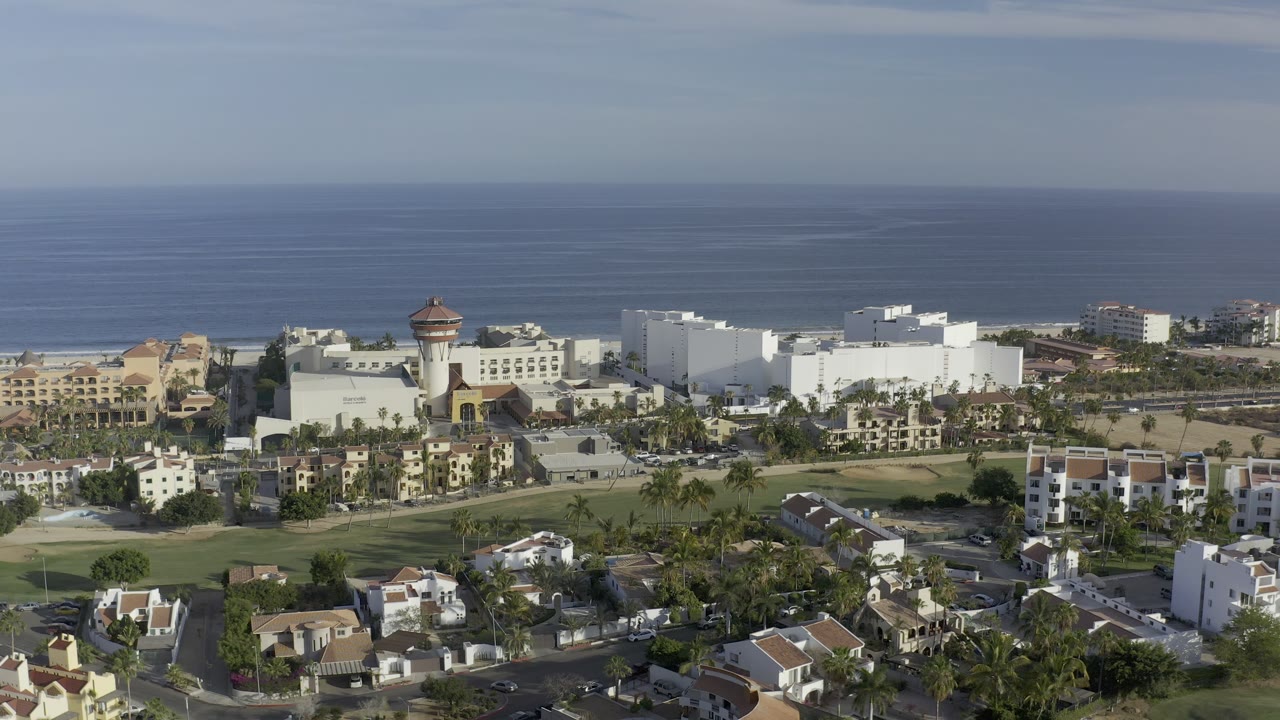 A Los Cabos coastal neighborhood fills the foreground, with palms and lawns around houses, large white buildings near the shoreline, and no visible people in the frame.