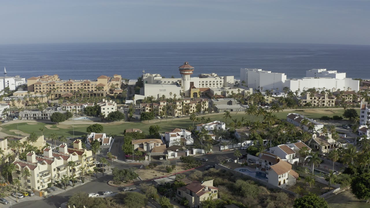 High aerial view over Los Cabos, Mexico, with tan and white coastal buildings, residential roofs, roads, palms, and a blue ocean horizon, no visible people.