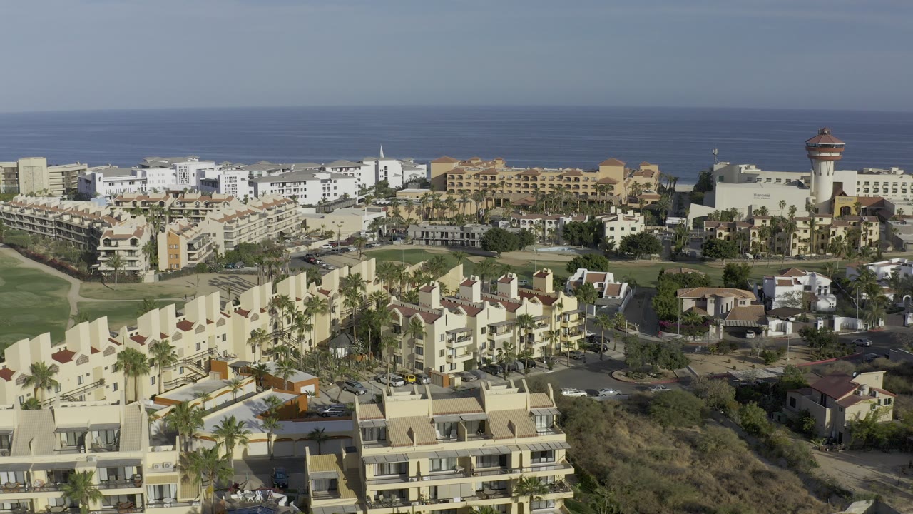 Aerial frame in Los Cabos showing cream vacation rental buildings, palm trees, green lawns, distant white structures, blue ocean horizon, and no visible people.