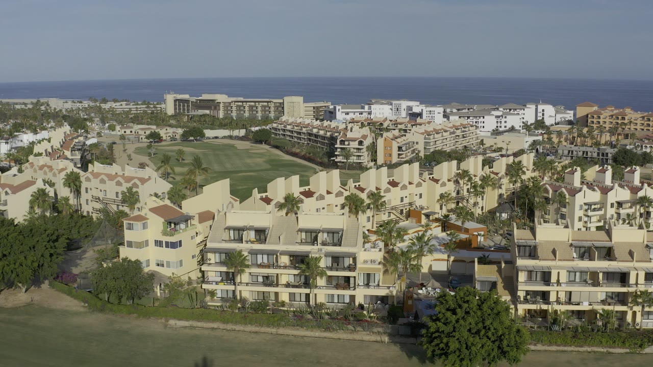 Elevated view of white buildings, green lawn, palms, and ocean horizon.