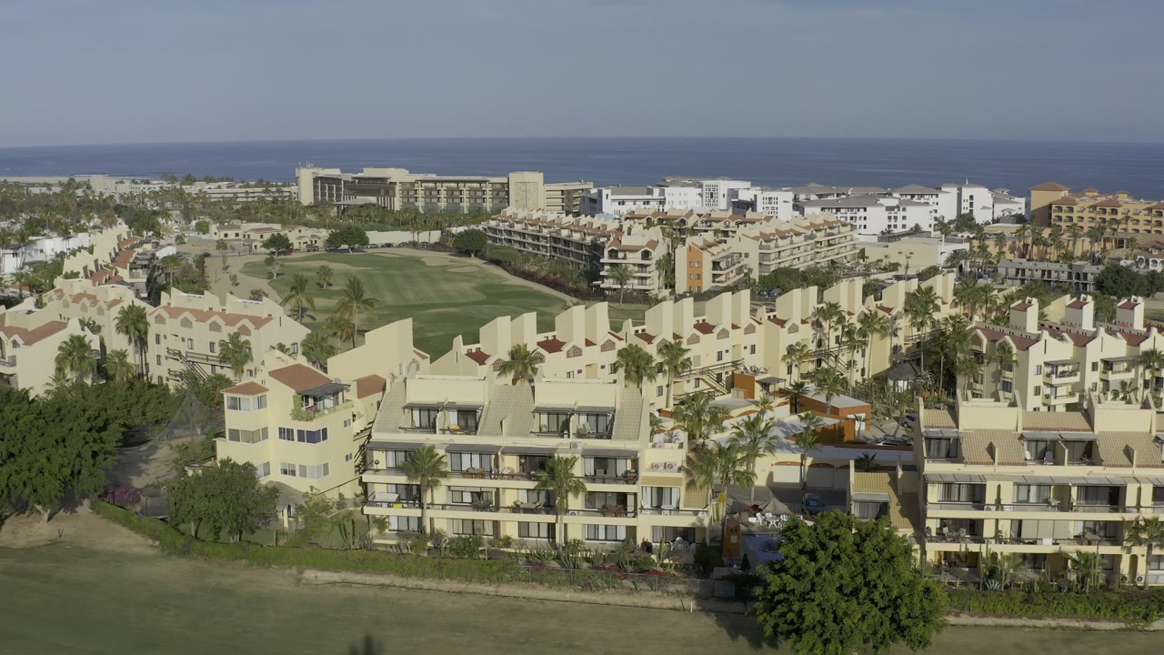 Los Cabos buildings with red-tile roof accents and balconies sit beside a broad lawn, with palm trees and the ocean line visible in the background, and no visible people.
