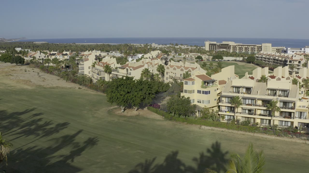 Early frame in Los Cabos, Mexico, looking across open grass toward pale houses, palms, dense trees, and a blue ocean horizon with no visible people.