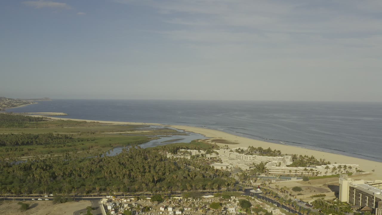 Wide aerial view over Los Cabos, Mexico, with a pale beach, blue ocean, green coastal vegetation, roads, and light buildings, with no visible people.