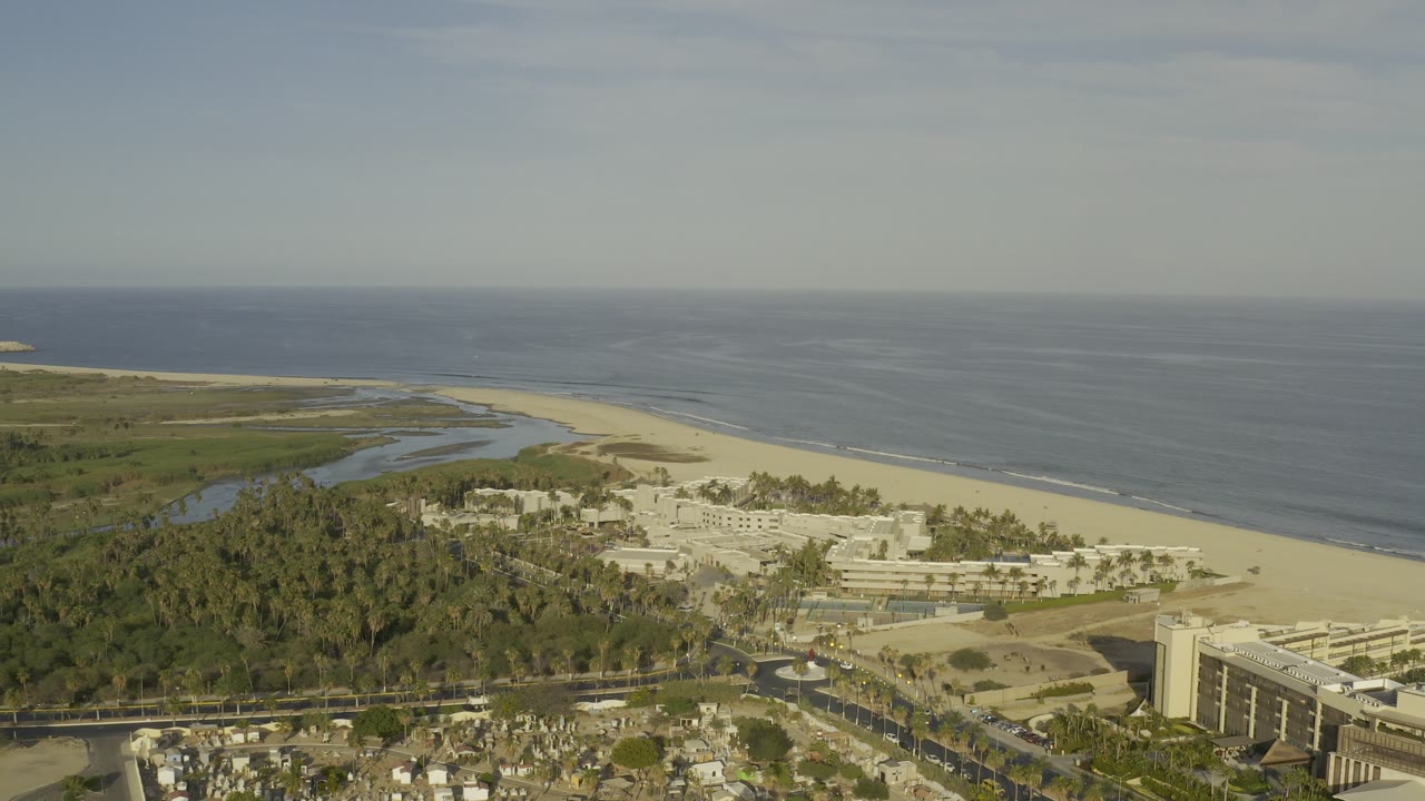 Aerial view over Los Cabos, Mexico, showing a sandy beach, dark blue ocean, green inlet, palms, and resort buildings with no visible people.