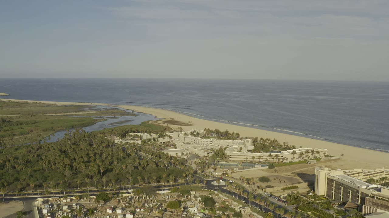Wide Los Cabos aerial frame with the ocean horizon across the top, a long sand beach, palm-covered land, white resort structures, and no visible people.