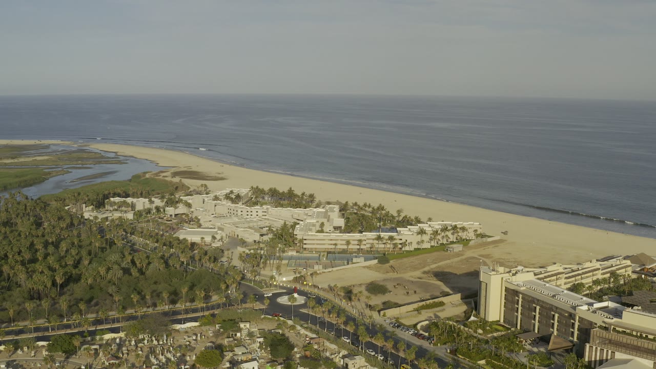Aerial view over Los Cabos, Mexico with a sandy beach, dark ocean, palm-covered grounds, resort-style buildings, and no visible people at the start of the clip.