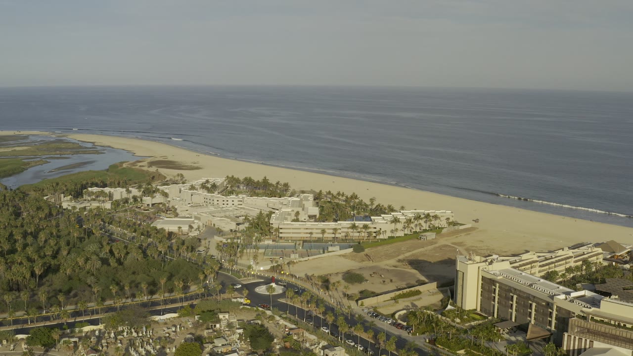 Drone-like view of ocean, beach, lagoon area, palms, and buildings.