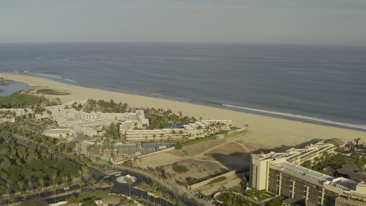 Aerial view over Los Cabos, Mexico with a pale beach, dark blue ocean, palm trees, roads, white coastal buildings, and no visible people.