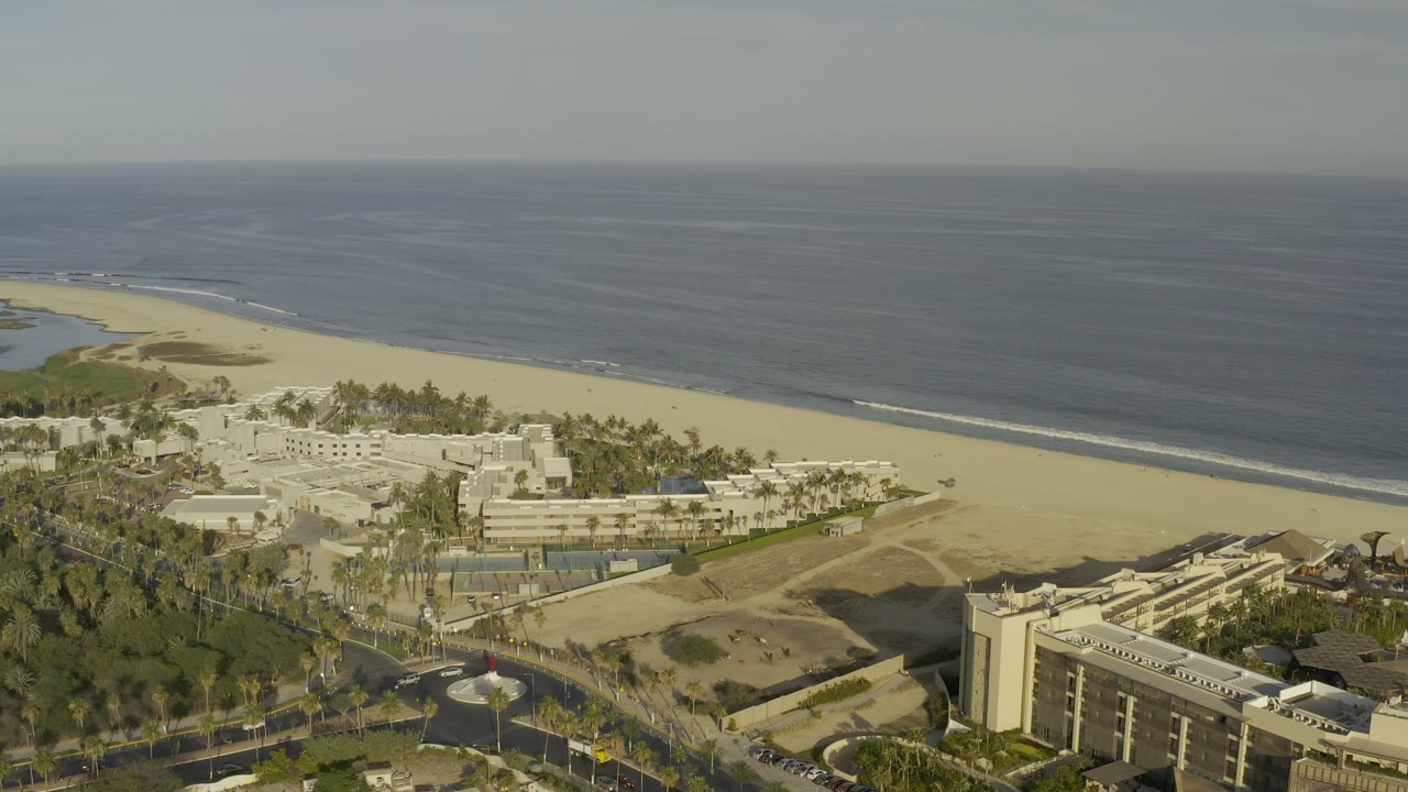High aerial frame in Los Cabos, Mexico showing the ocean horizon, wide sandy beach, palm-lined grounds, clustered white buildings, roads, and no visible people.
