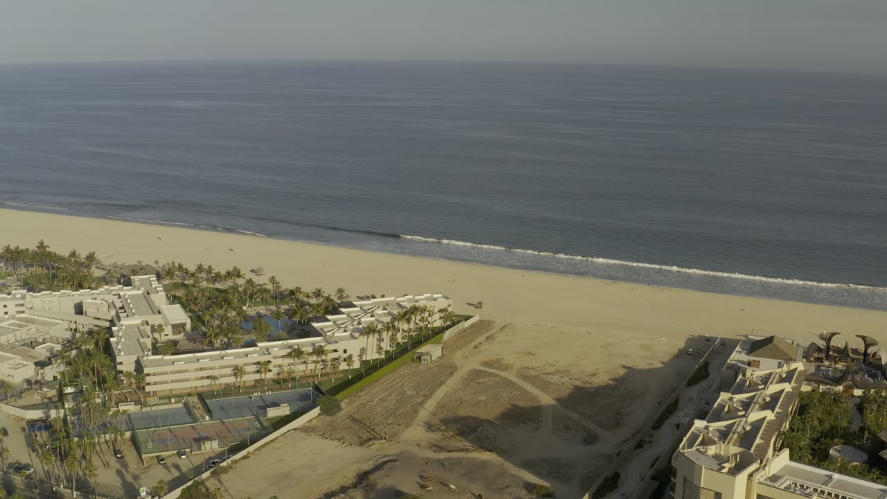Aerial Los Cabos shoreline view with pale beachfront buildings, palms, tennis courts, empty sand, a narrow surf band, and no visible people.