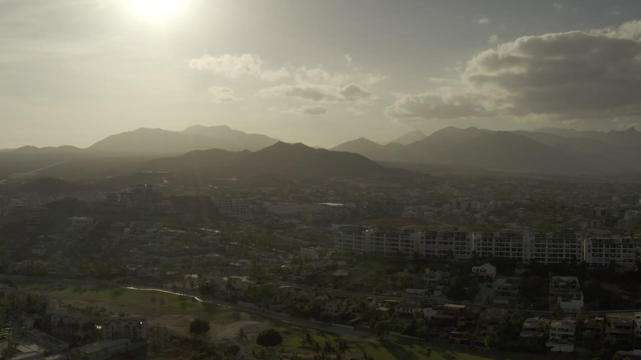 Wide aerial view over Los Cabos, Mexico, with city buildings below layered mountain ridges, bright sun near the top of frame, and no visible people.