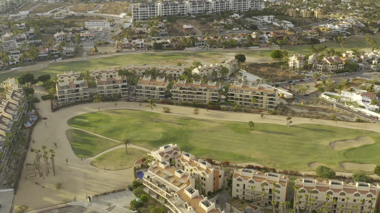 Aerial view in Los Cabos, Mexico showing tan resort buildings below a broad green courtyard area, sandy paths, roads, and distant structures, with no visible people.