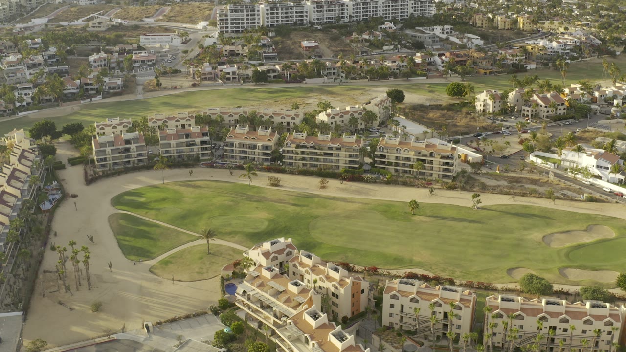 High oblique aerial in Los Cabos, Mexico with a manicured green courtyard, sand-colored open areas, resort blocks, roads, parked cars, and no visible people.