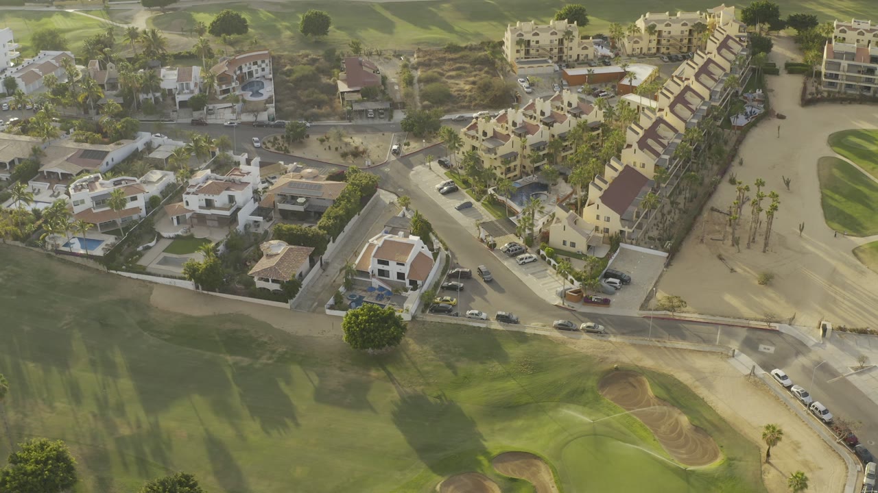 Aerial Los Cabos frame with tan-roofed buildings, white homes, parked cars along the road, a golf fairway in the foreground, and open sandy lots behind the houses.
