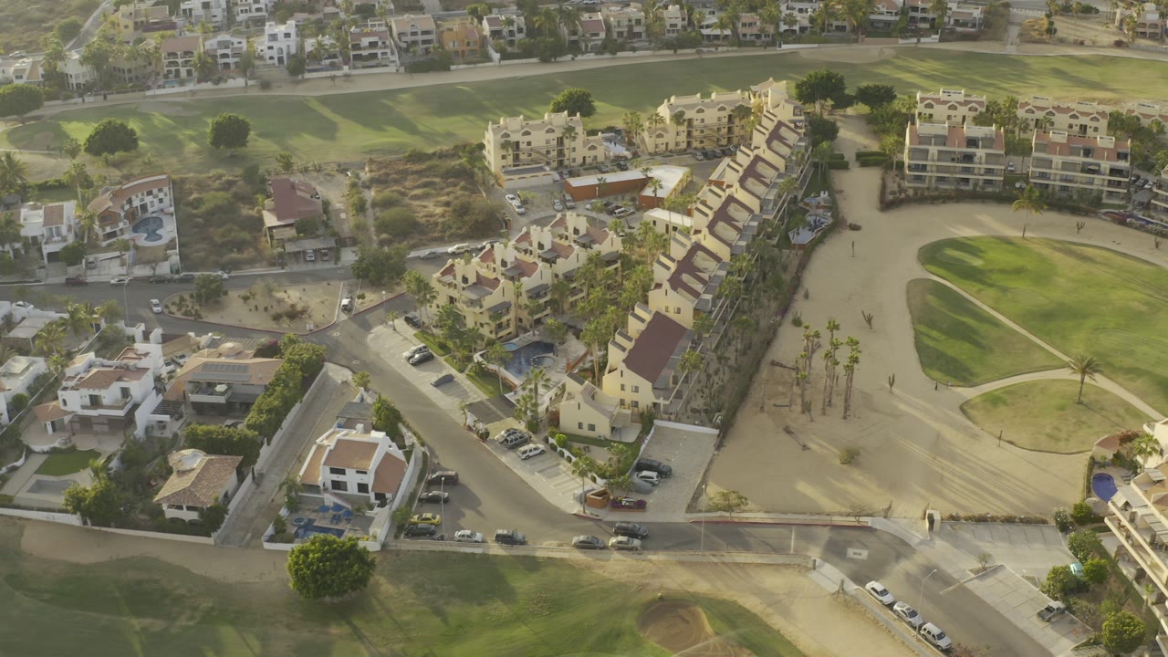 High aerial view in Los Cabos, Mexico, showing cream residential buildings, terracotta roofs, a green fairway, tan paths, roads, parked cars, and no visible people.