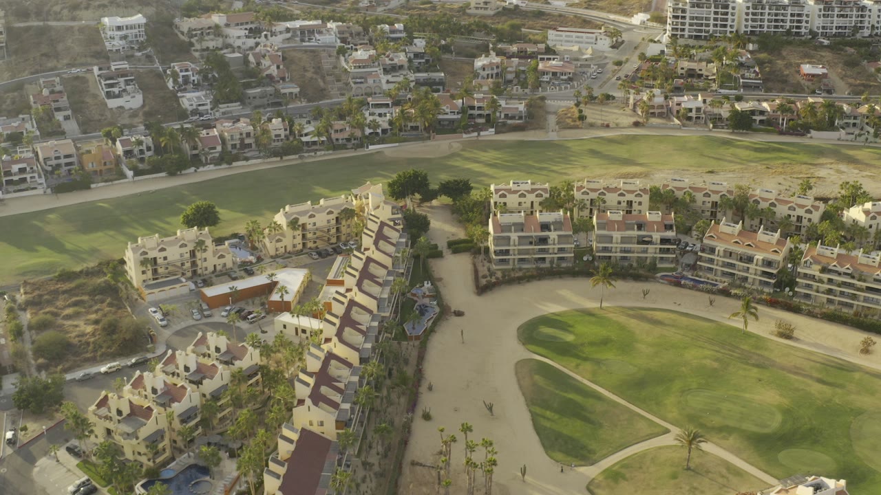 Drone view over Los Cabos, Mexico residences curving around a green landscaped area, with roads and hillside buildings in the background and no visible people.