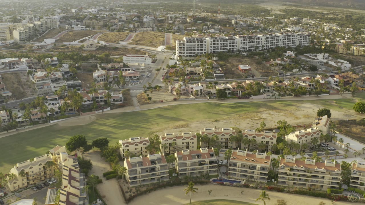 Opening aerial frame of Los Cabos, Mexico, showing cream residential buildings, a large green area, roads, dry terrain, distant hills, and no visible people.