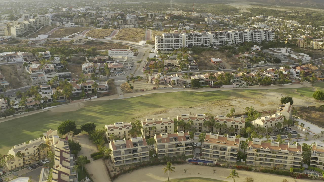 Aerial Los Cabos frame centered on white condominium blocks, neighborhood roads, a broad green open area, arid lots, hazy hills, and no visible people.