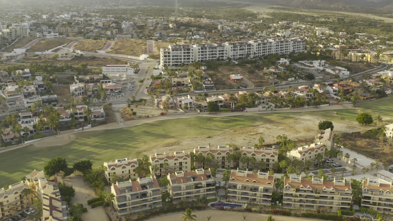 Aerial neighborhood view with clustered buildings, green course-like turf, roads, and hills.