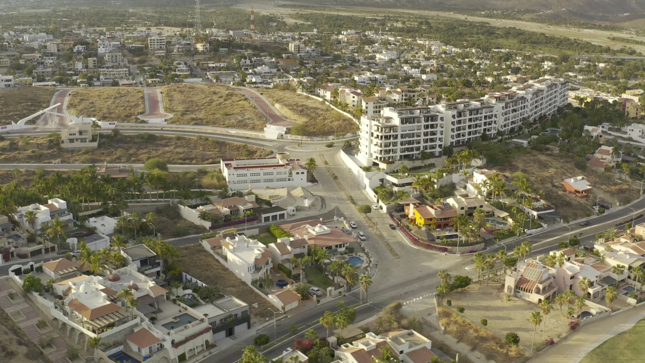 Elevated aerial frame over Los Cabos, Mexico, showing a road intersection, white residential buildings, dry lots, hillside development, and no visible people.