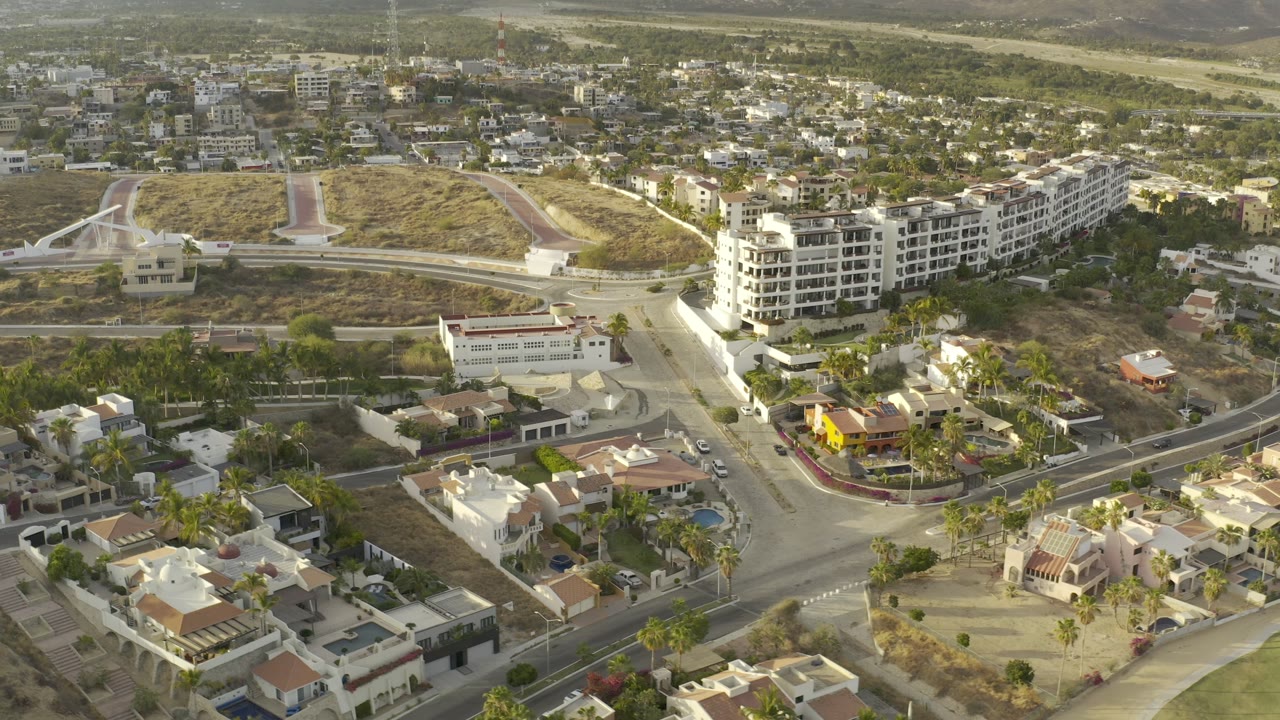 Wide aerial frame in Los Cabos, Mexico, with a white apartment block beside smaller homes, palm-lined lots, paved roads, dry hillside, and no visible people.