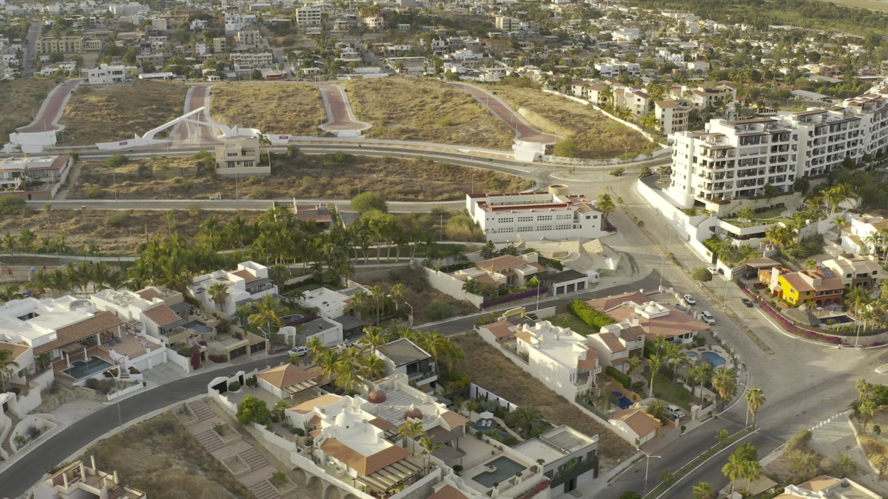 Elevated Los Cabos neighborhood frame showing white villas in the foreground, a road junction, dry lots, and apartment buildings beyond, with no clearly identifiable people visible.