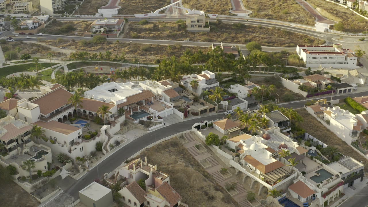 Aerial view of cream houses, terracotta roofs, palms, roads, and dry hillside.