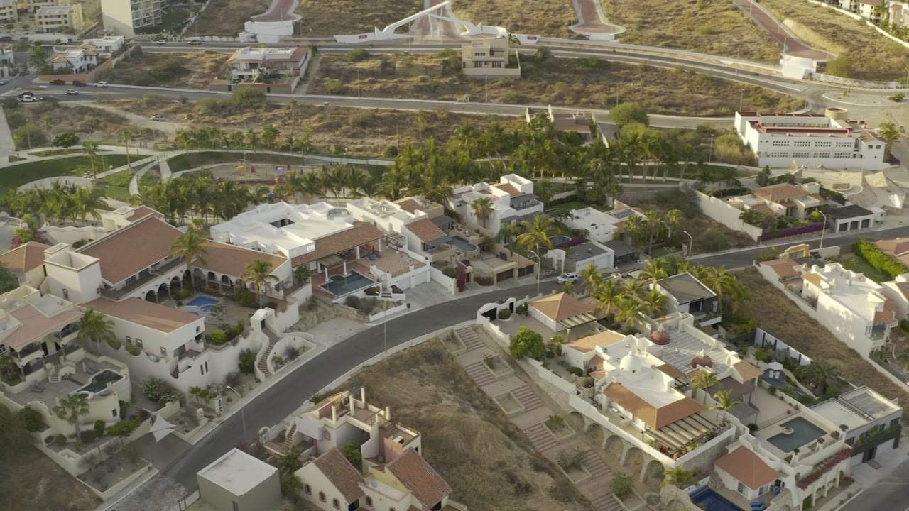 Aerial view in Los Cabos showing white and cream residential buildings, blue pools, palm landscaping, hillside streets, and no visible people.