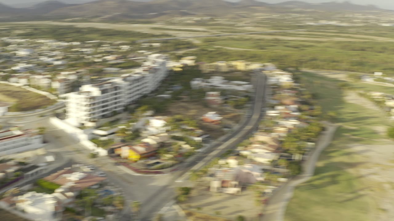 Opening aerial frame over Los Cabos, Mexico, with soft-focus buildings, curved roads, inland hills, green ground, and no visible people.