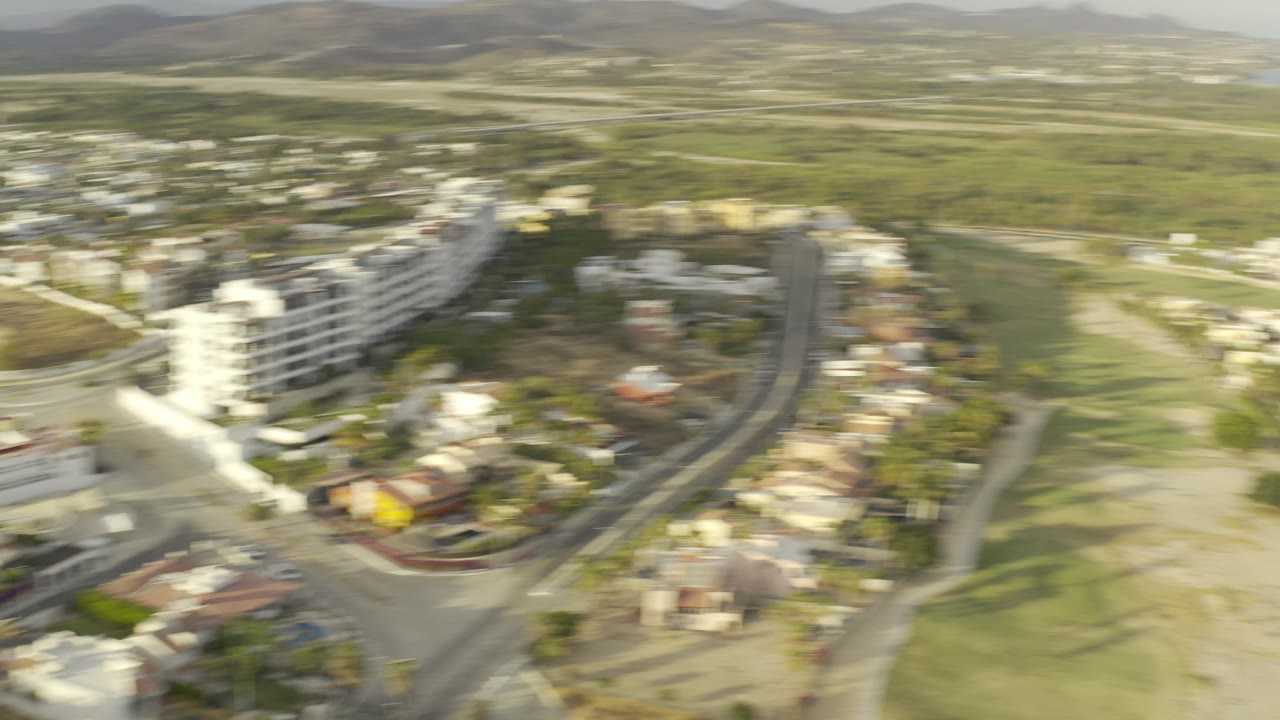 Second aerial frame in Los Cabos shows white multi-story buildings, nearby roads, green open land, distant hills, and no visible people.