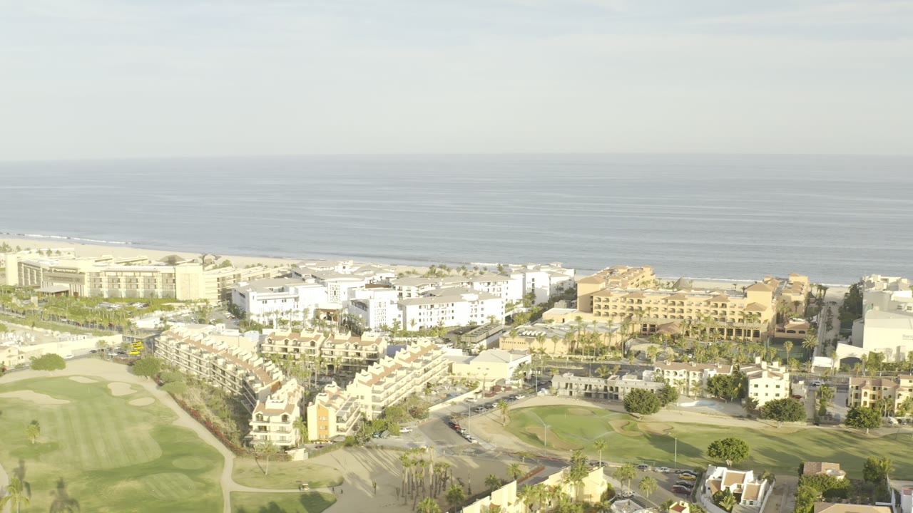 Aerial view in Los Cabos, Mexico, with rows of white residential buildings, tan structures, palm trees, roads, golf greens, and open sea in the background, with no visible people.