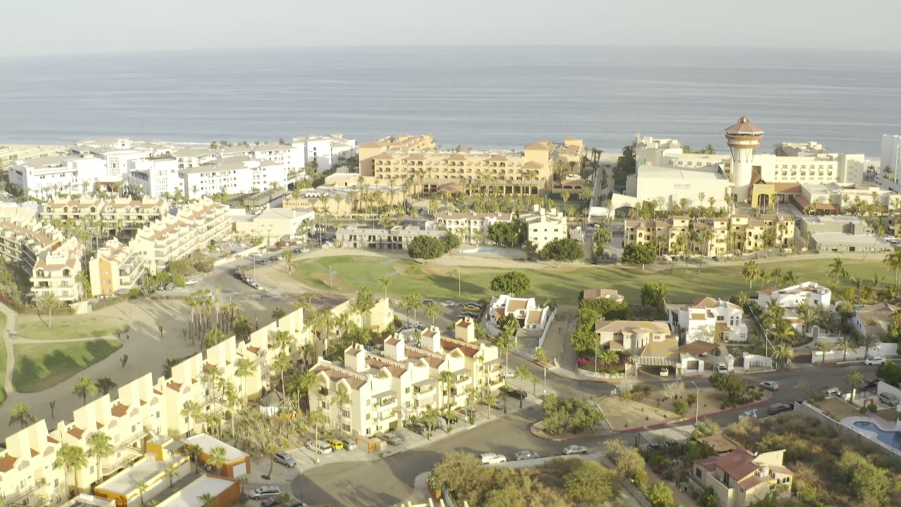 Aerial frame over Los Cabos, Mexico showing villas and palm-lined streets in the foreground with pale resort buildings and the sea beyond, no visible people.
