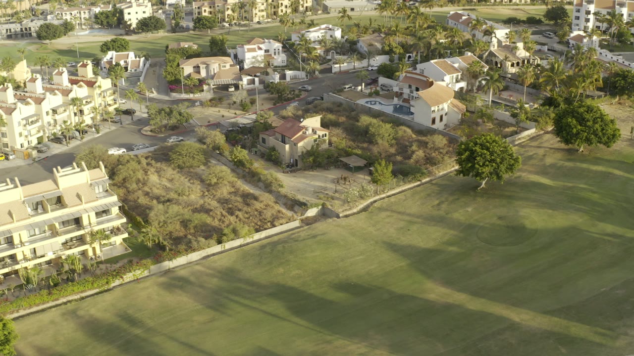 A high aerial frame over Los Cabos shows green fairway grass, low-rise homes, palms, dry brush, and no visible people.
