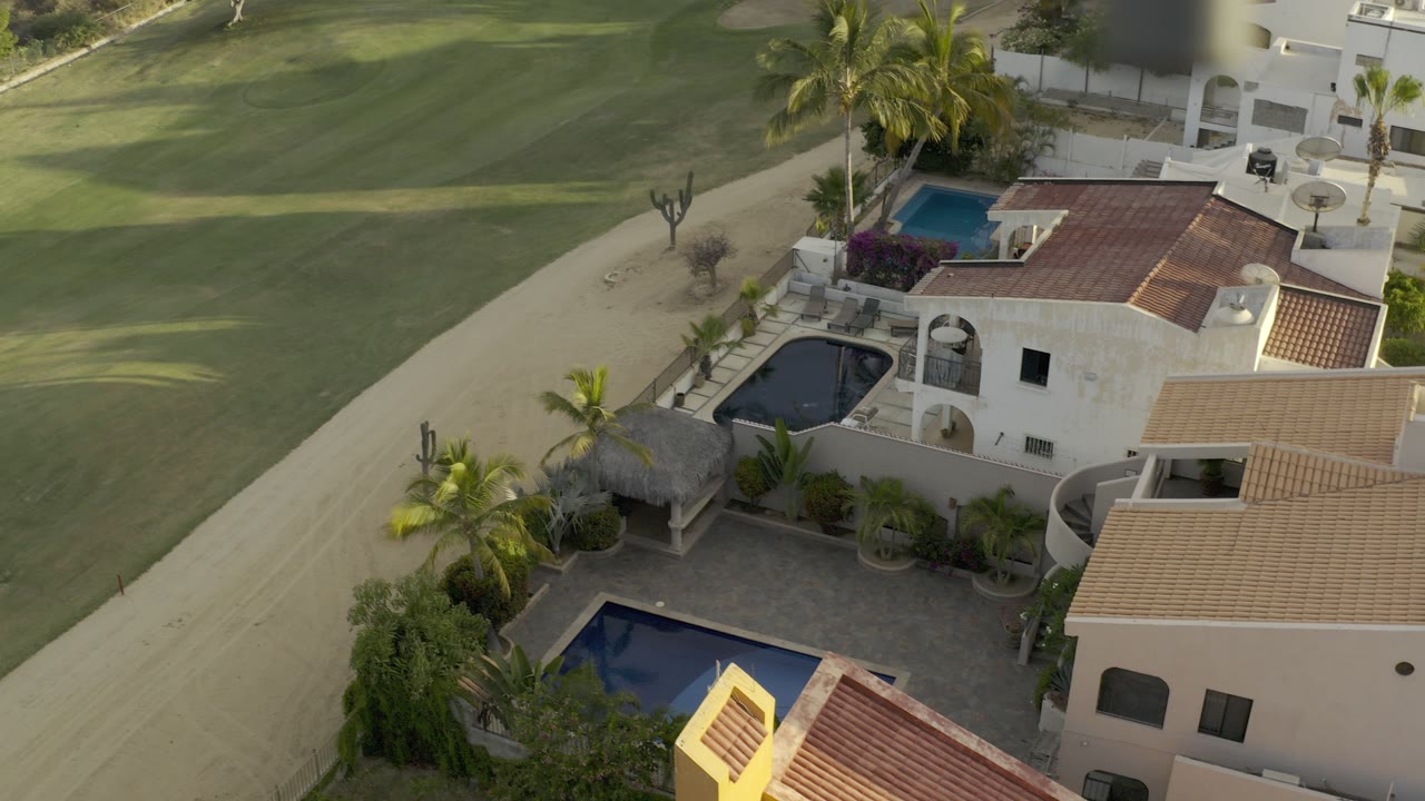 Aerial view of tile-roof houses, pools, palms, and a sandy road.