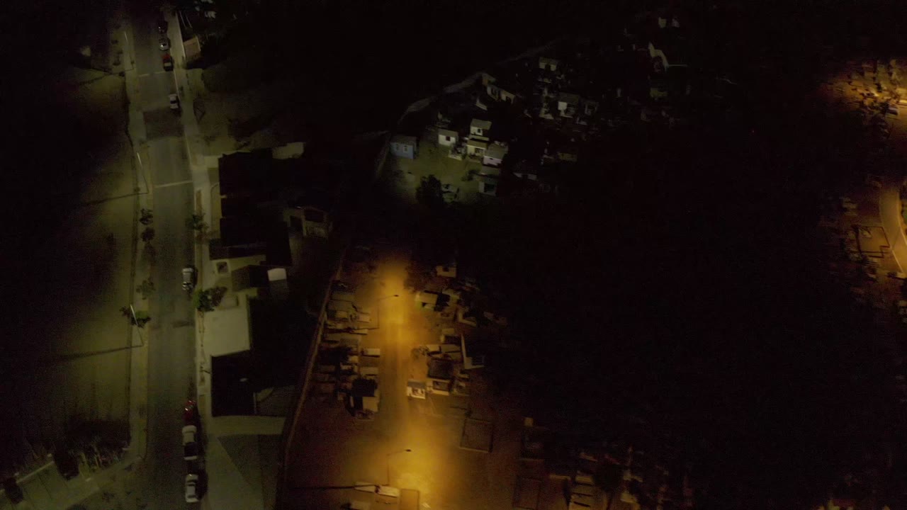 Los Cabos night aerial showing the road shifted slightly left in frame, a warm amber-lit area near rooftops, parked vehicles, and no visible people.