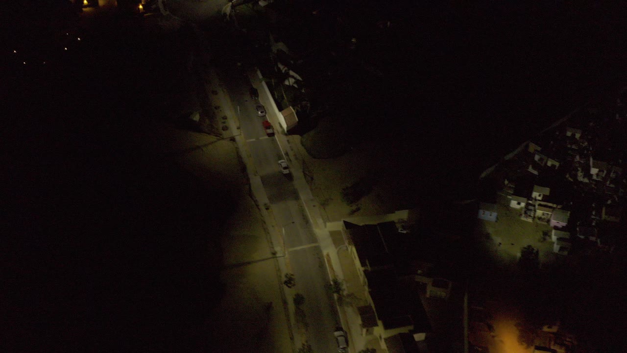 Night aerial frame over Los Cabos, Mexico showing an angled residential street, warm streetlights, dark surrounding areas, parked cars, and no visible people.