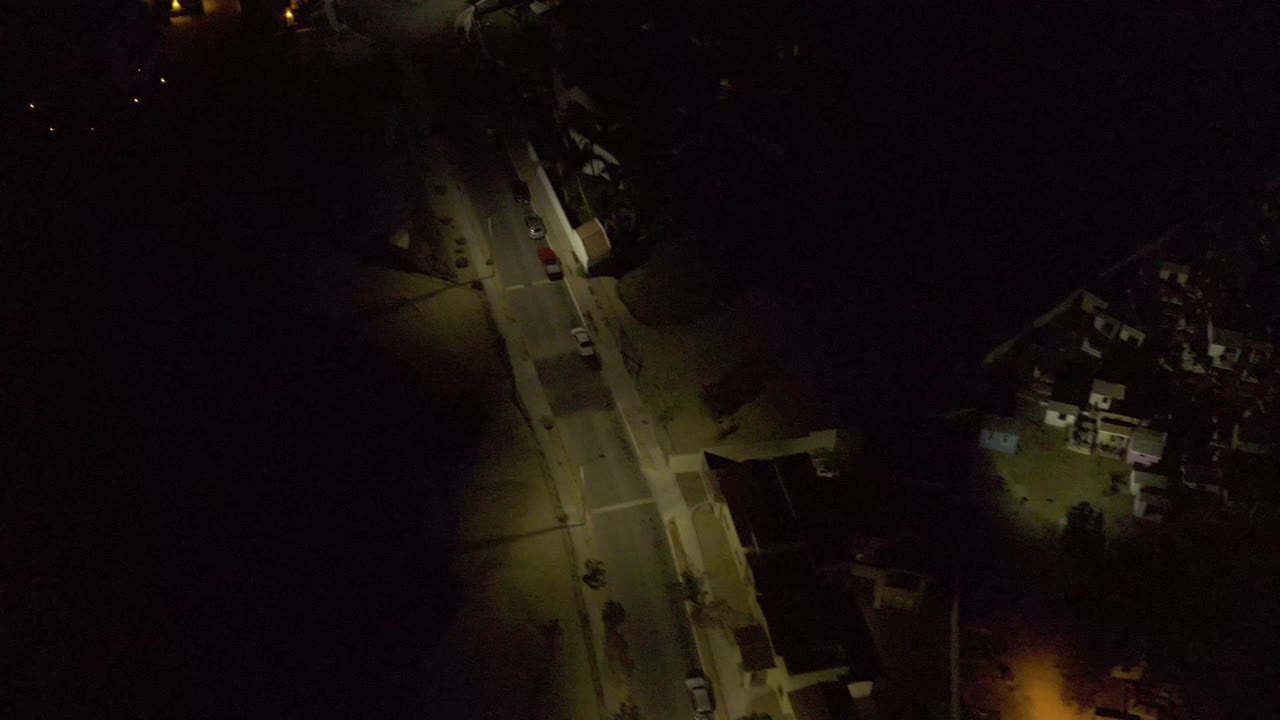 Night drone view in Los Cabos, Mexico with the road slightly farther through frame, lit sidewalks, clustered homes, parked cars, and no visible people.