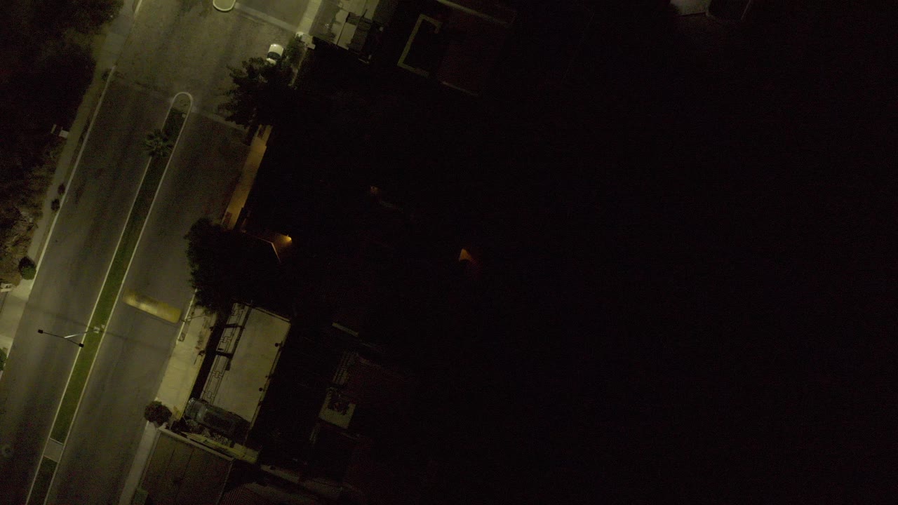 Overhead night view in Los Cabos, Mexico, showing a dim street, narrow sidewalk, trees, and dark building edges with no visible people.