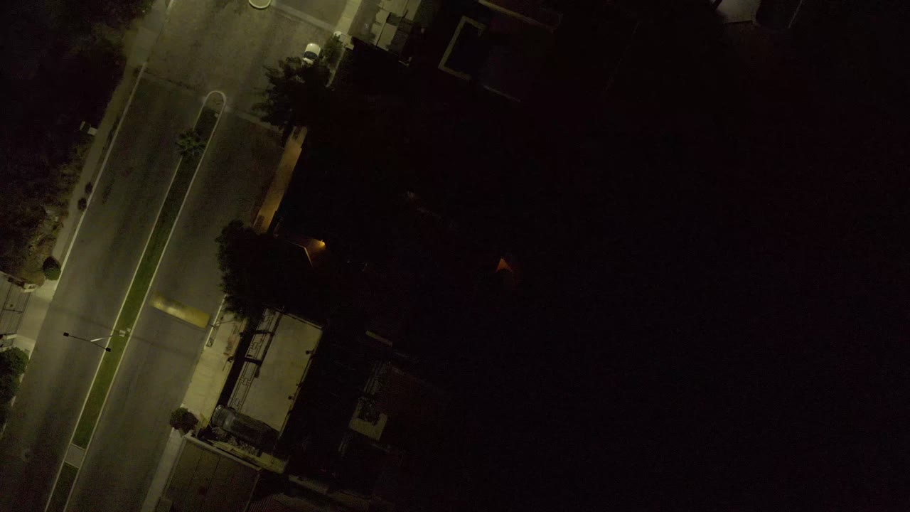 Night aerial frame in Los Cabos, Mexico, showing the road edge, a small illuminated roadside building, deep tree shadows, and no visible people.