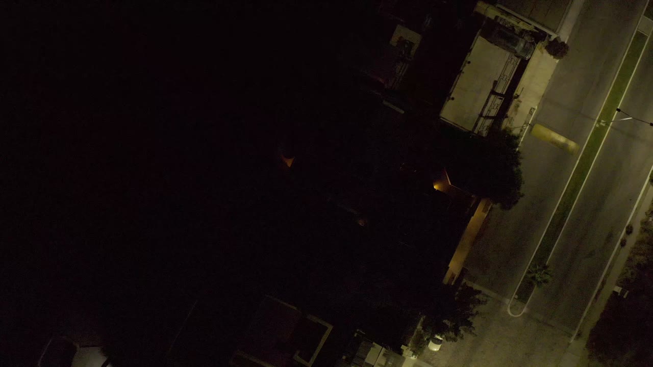 Aerial frame in Los Cabos, Mexico, with the empty road cutting diagonally past a lit curb, dark trees, and a small building edge, with no visible people.