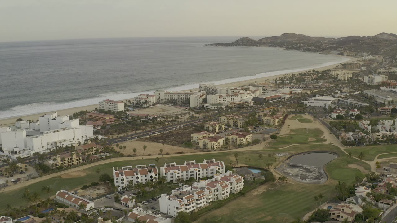 Aerial view of ocean, beach, coastal buildings, lawns, and distant hills.