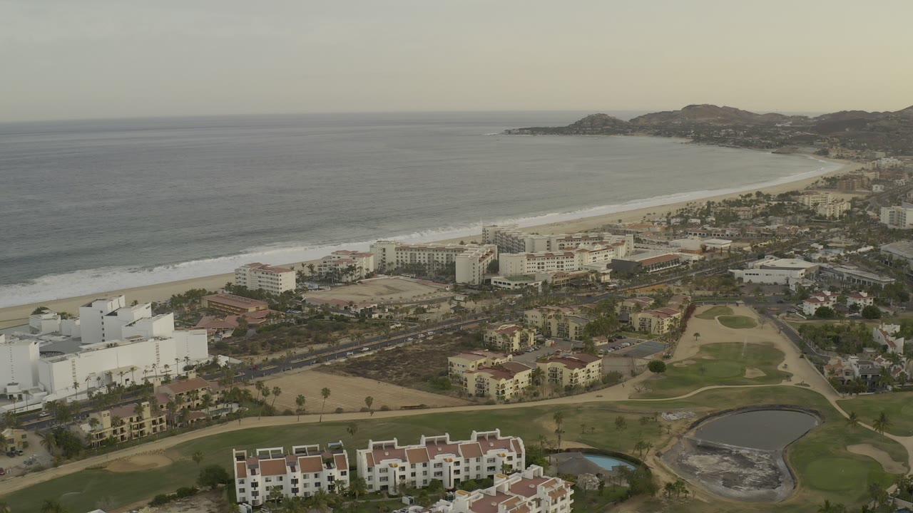 High aerial view over Los Cabos, Mexico, with hotels near a sandy beach, surf lines, green grounds, ponds, distant hills, and no visible people.