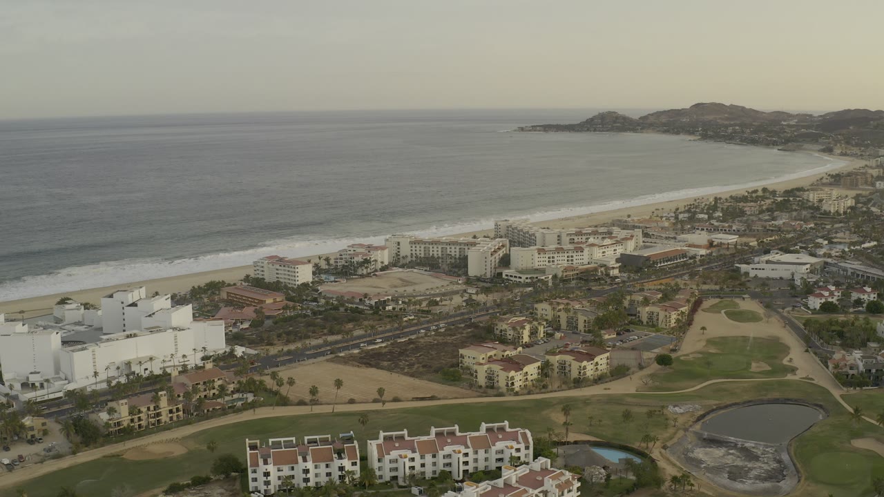 Drone view of coastal hotels beside surf and a sandy beach.