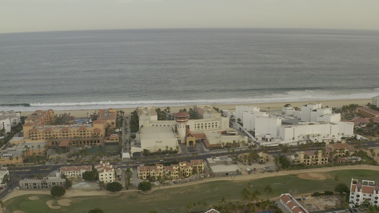Aerial view of Los Cabos, Mexico with ocean surf beyond beachfront hotels, roads, golf course greens, and no visible people.