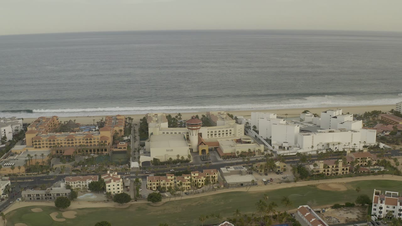 Los Cabos aerial frame with a broad gray-blue ocean horizon, beachfront hotel complexes, a road grid, golf course fairways, and no visible people.