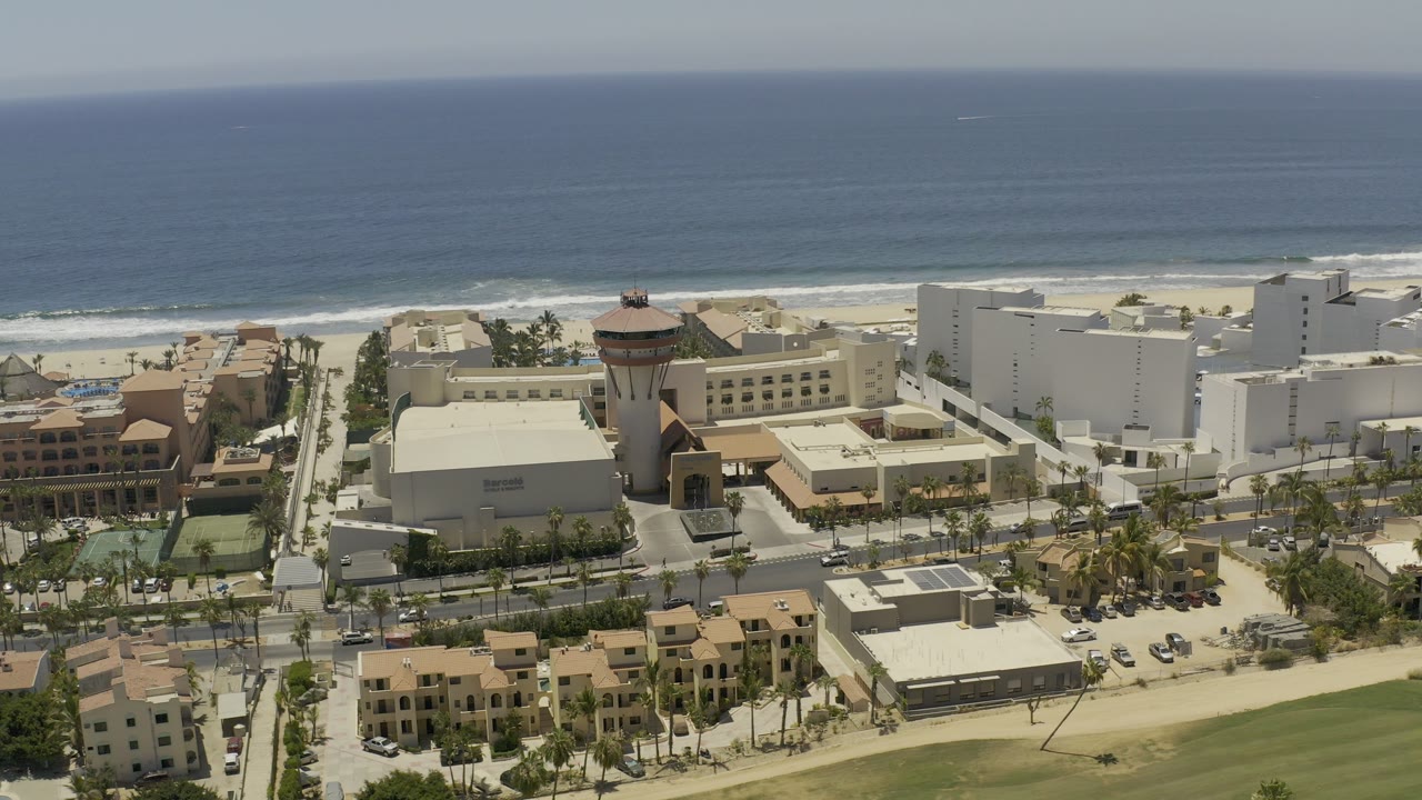 Opening aerial frame over Los Cabos, Mexico, with the ocean horizon above beachfront buildings, palm-lined roads, parked cars, and no visible people.