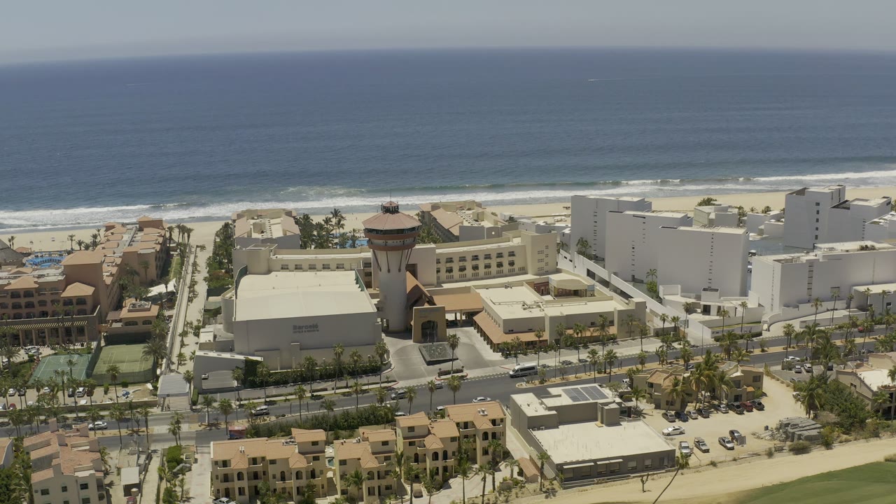 High aerial frame over Los Cabos beachfront buildings and roads, with the ocean horizon higher in the frame and no visible people.