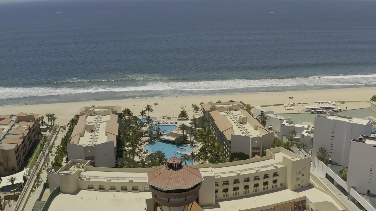 High aerial frame in Los Cabos, Mexico showing the resort pool area slightly right of center, hotel roofs below, a quiet beach strip, and parallel white wave lines offshore.